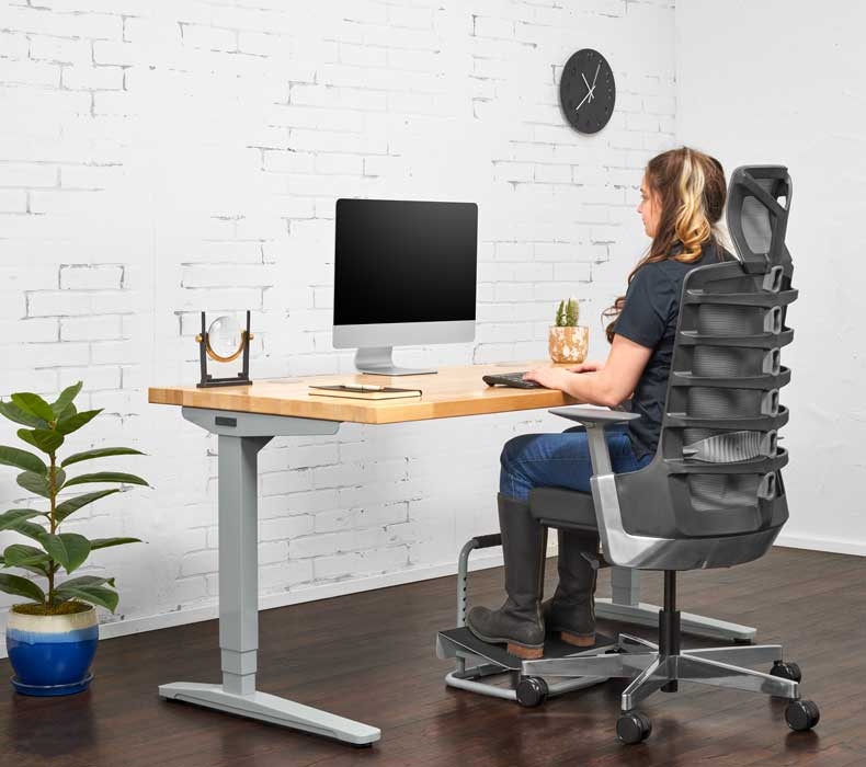 A person sits at a height adjustable desk and raises their feet with an office footrest