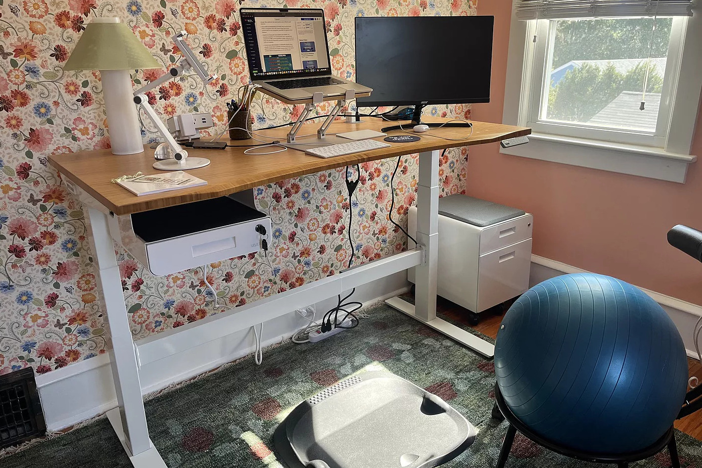 A colorful home office with floral wallpaper, featuring an Uplift Standing Desk with an Eco-Curve Bamboo desktop.