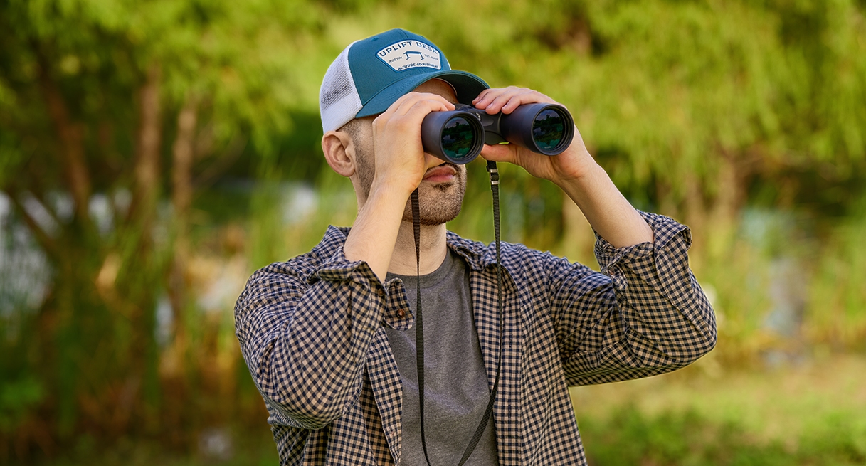 Man looking through binoculars outside while wearing an UPLIFT Desk Cap