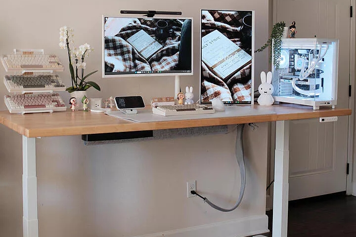 Alt: Uplift Standing Desk with a solid wood desktop in a cozy office setting with multiple monitors.
