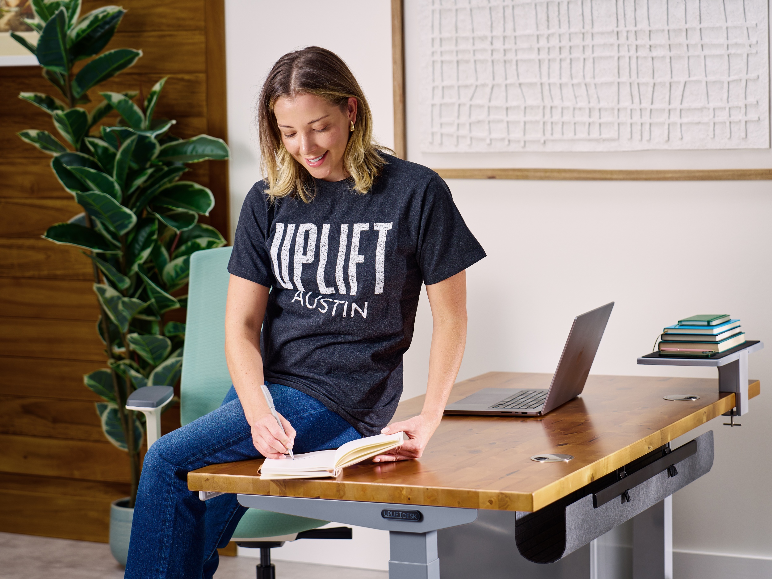 A woman sits on top of a V3 2-leg standing desk and writes in a notebook