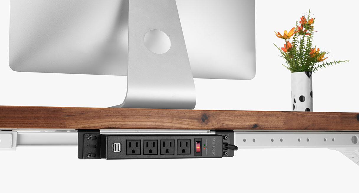A 4-outlet power strip mounted to the underside of a desk using desktop brackets with the outlets facing the rear