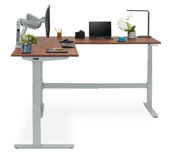 L-Shaped Standing Desk with walnut desktop on a gray height adjustable frame featuring a Crestview Dual Monitor Arm in gray and desk accessories.